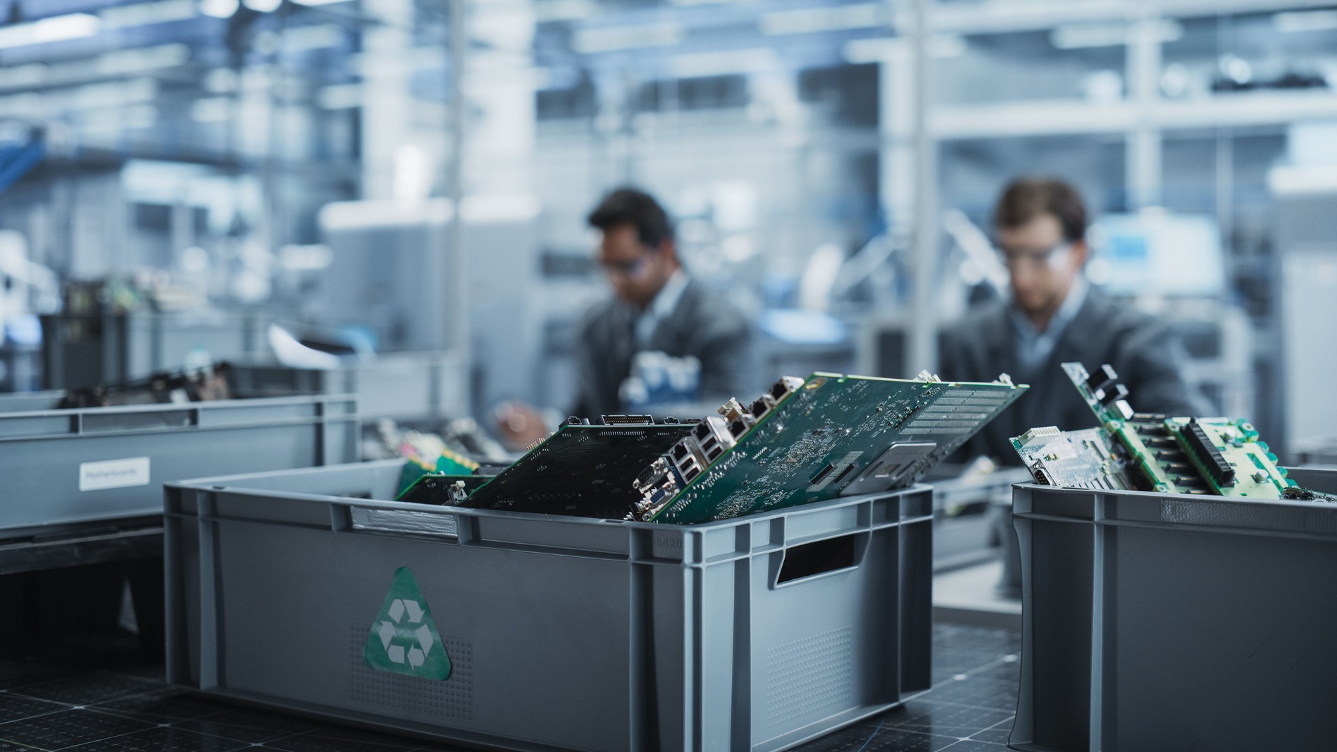 Indian And Caucasian Male Workers Taking Apart Laptops To Recycle For Production Of Digital Devices At Automated Electronics Factory With Robotic Arms. Colleagues Sorting Displays, Keyboards.